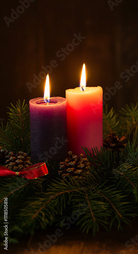 Two lit candles, one purple and one red, surrounded by pine branches, small pinecones, and a red ribbon.