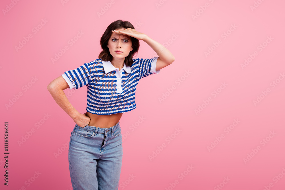 Young woman in striped knitwear top looking ahead with hand shielding eyes against pink background Young woman in striped knitwear top looking ahead with hand shielding eyes against pink background