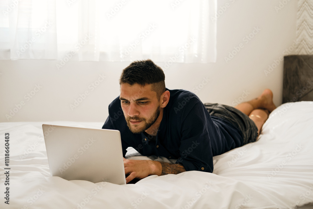 A young Caucasian bearded man is lying on a bed with a laptop. Working online from home concept. The guy is a freelancer at home interior A young Caucasian bearded man is lying on a bed with a laptop. Working online from home concept. The guy is a freelancer at home interior