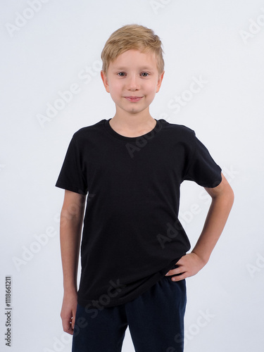 Pleased blonde haired boy points at blank black t shirt shows place for your advertisement focused down isolated over white background. People clothing and promotion concept.