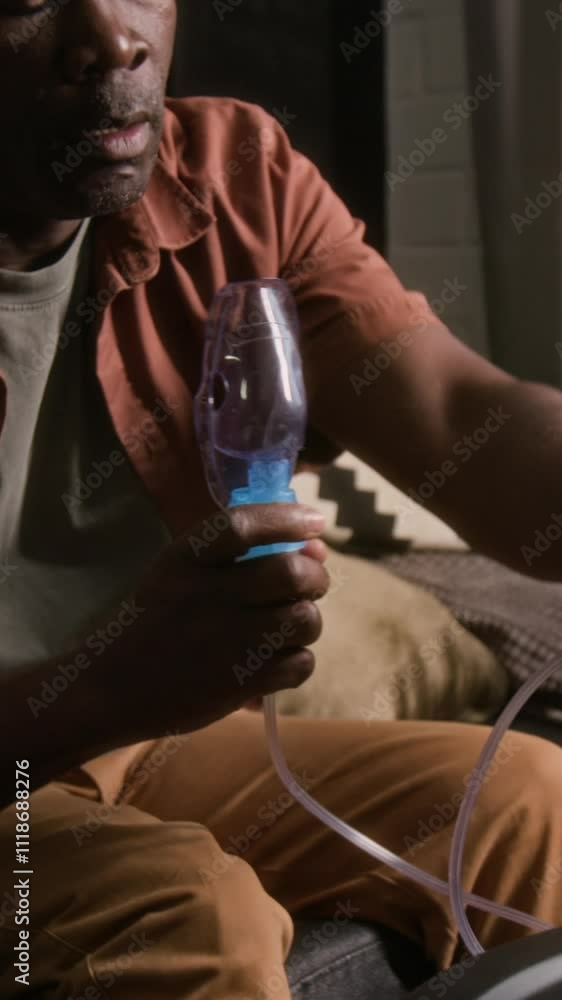 Vertical shot of mature African American man using compressor nebulizer mask, leaning back on couch and scrolling on smartphone during asthma treatment at home Vertical shot of mature African American man using compressor nebulizer mask, leaning back on couch and scrolling on smartphone during asthma treatment at home