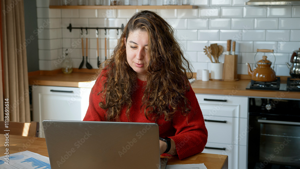 A woman works from home, seated at a table in a modern kitchen. Holding a document while a laptop is open in front of her. A woman works from home, seated at a table in a modern kitchen. Holding a document while a laptop is open in front of her.