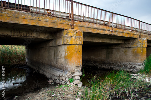 A deteriorating bridge with rusty railings and cracked concrete pillars, spanning a shallow waterway.
