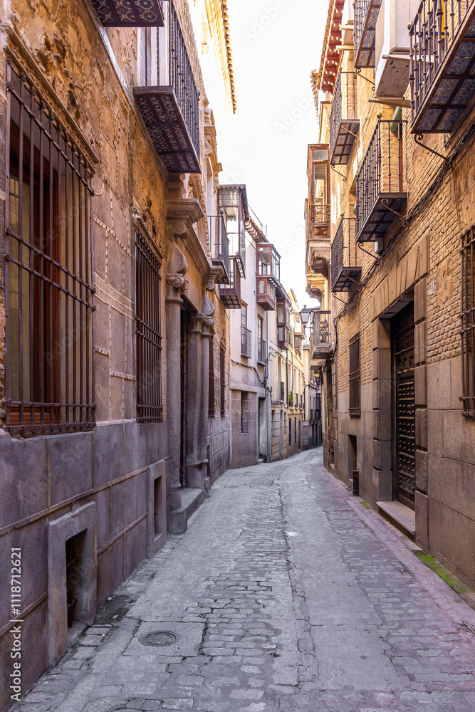 Fototapeta premium Narrow Spanish medieval cobbled street with old residential houses, traditional forged balconies and old-style street lamps, Toledo, Spain.