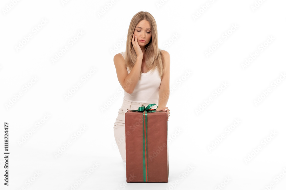 A young woman looks puzzled while kneeling beside a wrapped gift in a bright studio setting A young woman looks puzzled while kneeling beside a wrapped gift in a bright studio setting