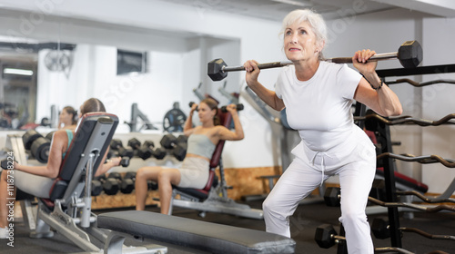 Slender senior woman trains muscles of arms and back using barbell and differentiated weighting in crossfit gym. Lunges and squats with additional equipment and barbell weight