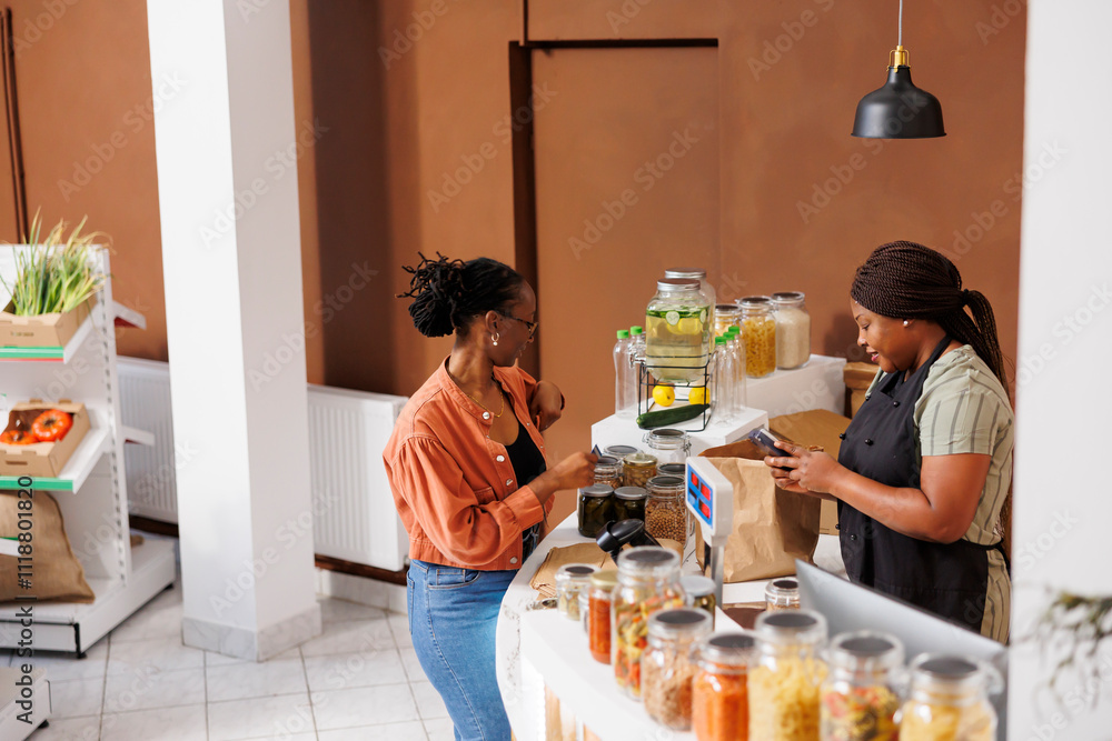 © DC Studio - Black woman returning card into pocket after paying for bio food products and items she chose at an environmentally friendly store. Vendor and customer completing cashless transaction at store counter © DC Studio - Black woman returning card into pocket after paying for bio food products and items she chose at an environmentally friendly store. Vendor and customer completing cashless transaction at store counter
