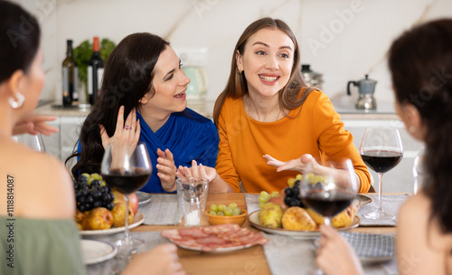 Cheerful female friends enjoying home gathering with wine and light snacks at kitchen table, engaging in lively conversations..