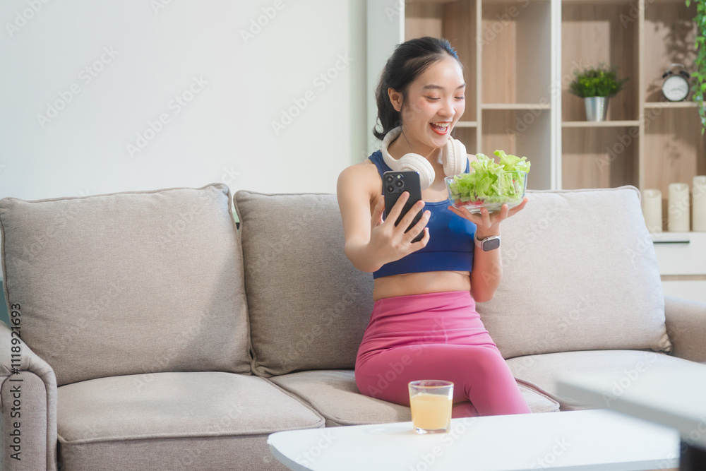 A young Asian housewife enjoys a healthy lifestyle at home, balancing fitness routines with relaxation. She sits on a modern sofa, eating a fresh salad and fruit while embracing healthy habits. A young Asian housewife enjoys a healthy lifestyle at home, balancing fitness routines with relaxation. She sits on a modern sofa, eating a fresh salad and fruit while embracing healthy habits.