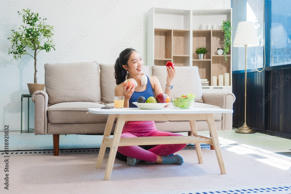 A young Asian housewife enjoys a healthy lifestyle at home, balancing fitness routines with relaxation. She sits on a modern sofa, eating a fresh salad and fruit while embracing healthy habits. A young Asian housewife enjoys a healthy lifestyle at home, balancing fitness routines with relaxation. She sits on a modern sofa, eating a fresh salad and fruit while embracing healthy habits.
