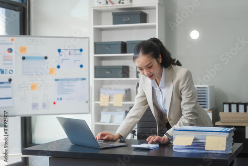 Fotografía A young Asian businesswoman works alone at a desk with a laptop, stack of papers, and documents