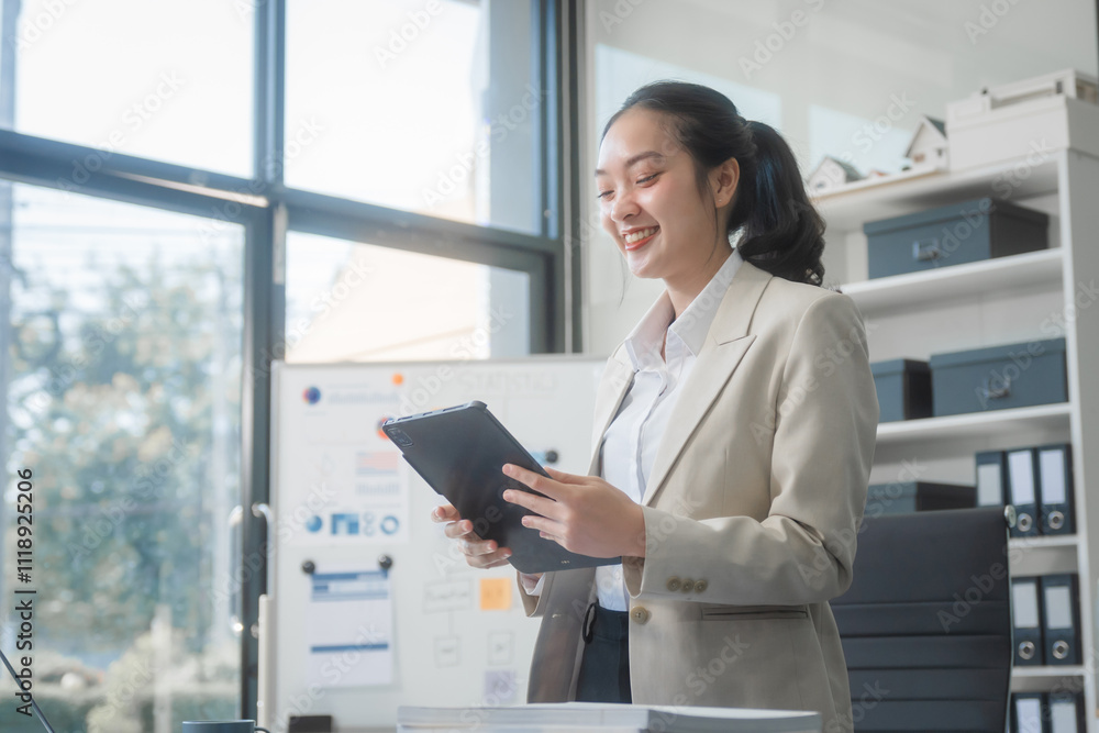 A young Asian businesswoman works alone at a desk with a laptop, stack of papers, and documents. She looks happy and successful, managing accounts and preparing financial statements in daylight. A young Asian businesswoman works alone at a desk with a laptop, stack of papers, and documents. She looks happy and successful, managing accounts and preparing financial statements in daylight.