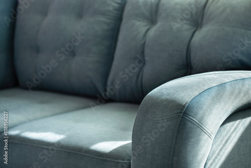 Soft blue velour sofa with quilted cushions and stitched armrest in shadows and sunlight. Part of the sofa with a soft back and seat close-up with copy space.
