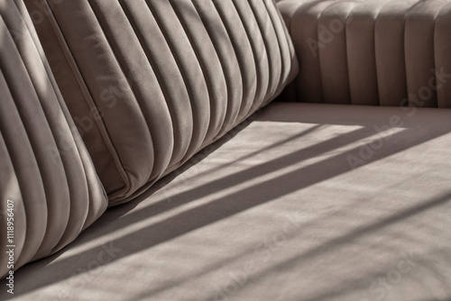 Soft beige velour sofa quilted with straight lines of stitching in shadows and sunlight. Part of the sofa with armrest and pillows close-up with copy space.