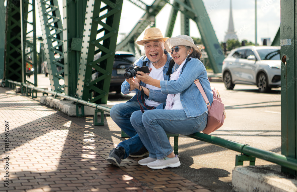 An elderly couple relaxes and looks at photos in camera after walking through the city streets with the road and bridge structures in the background An elderly couple relaxes and looks at photos in camera after walking through the city streets with the road and bridge structures in the background