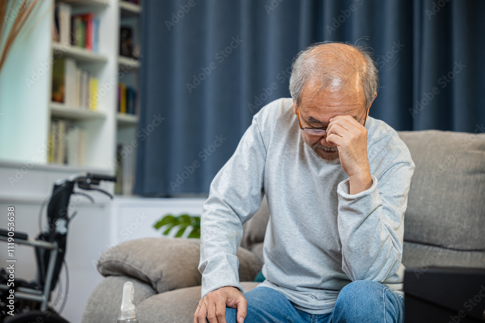 Fototapeta premium Asian mature old man sitting on sofa headache at home in living room, Depressed senior man unhappy hands rubbing eye suffering from a migraine. Health care