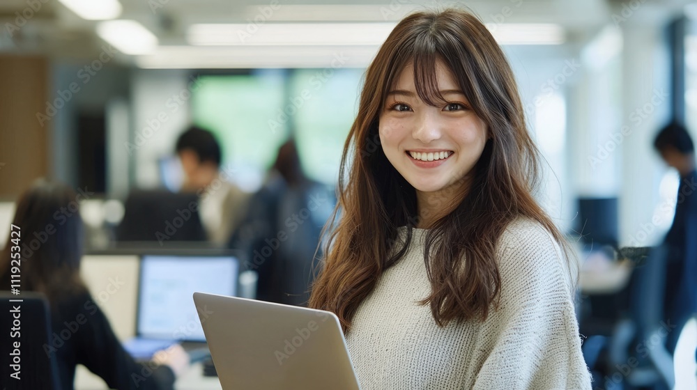 © Ganesha88 - Smiling Japanese businesswoman standing in an office, holding her laptop. She is wearing casual clothes, and other people working behind her are blurred.,generated ai.