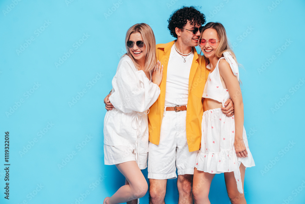 Group of young three stylish friends posing in studio. Fashion man and two cute female dressed in casual summer clothes. Smiling models having fun. Cheerful women and guy near blue wall Group of young three stylish friends posing in studio. Fashion man and two cute female dressed in casual summer clothes. Smiling models having fun. Cheerful women and guy near blue wall