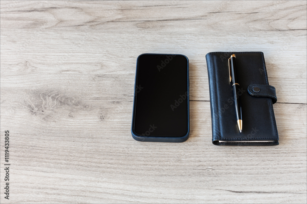 There is a closed black notebook, a fountain pen and a smartphone on the wooden tabletop. There is a closed black notebook, a fountain pen and a smartphone on the wooden tabletop.