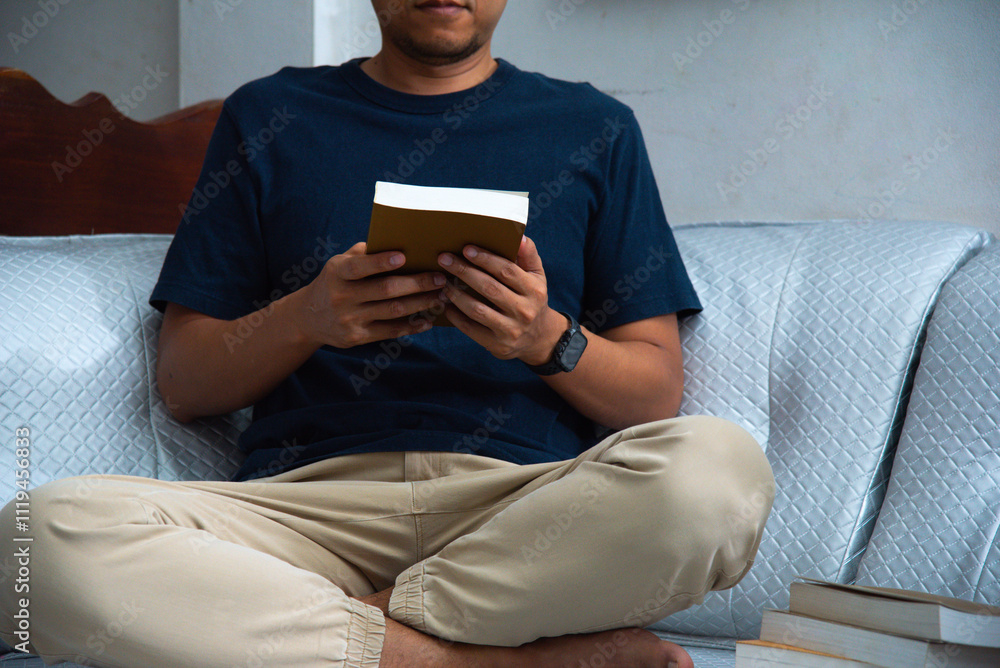 A man is seated comfortably on a sofa, engaged in reading a book. His attire is casual yet neat, such as a shirt and trousers, suggesting a moment of leisure within a serene environment. A man is seated comfortably on a sofa, engaged in reading a book. His attire is casual yet neat, such as a shirt and trousers, suggesting a moment of leisure within a serene environment.