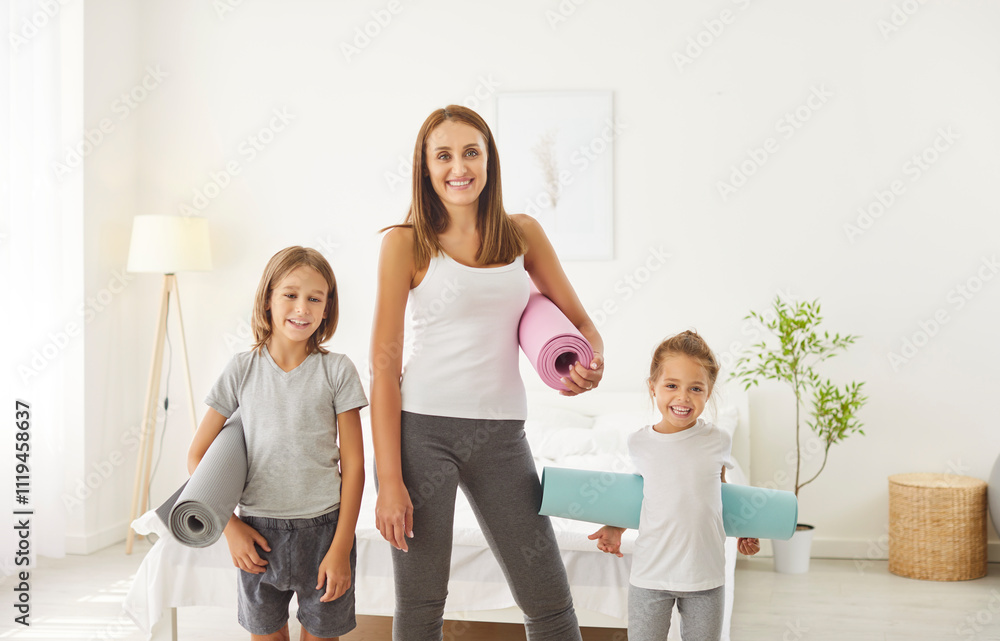 Fototapeta premium Happy woman in sport clothes standing in bedroom with her children, preparing to home workout together. Cheerful sporty mother and kids holding yoga mats and looking at camera. Healthy family concept.