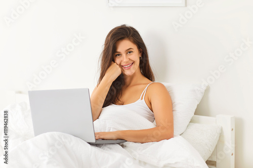 Relaxed happy smiling woman in bed with laptop, working online, spending carefree day, chatting on social media, making remote telecommuting in cozy home, using online services, funny morning start