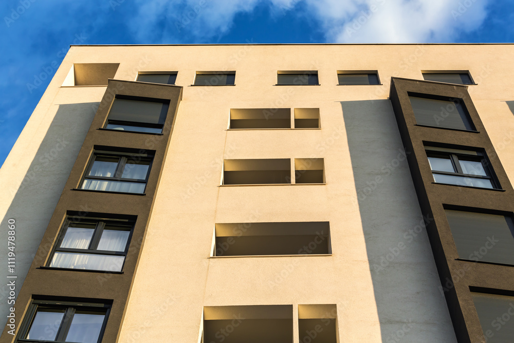 Modern apartment building facade with contrasting beige and dark panels, geometric windows, and natural light under a bright blue sky Modern apartment building facade with contrasting beige and dark panels, geometric windows, and natural light under a bright blue sky