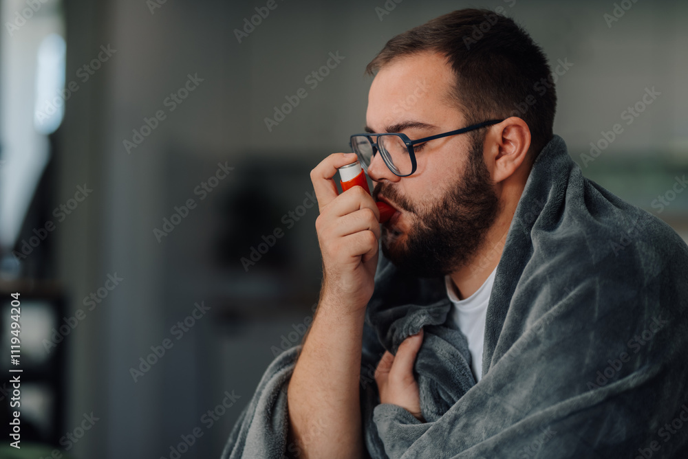 Sick man using inhaler at home wrapped in blanket Sick man using inhaler at home wrapped in blanket
