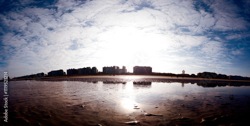 A tranquil scene of a coastal city with skyscraper silhouettes and tranquil water reflections under a vast, cloud-filled sky. The sunlight creates a serene atmosphere on the sandy shoreline.