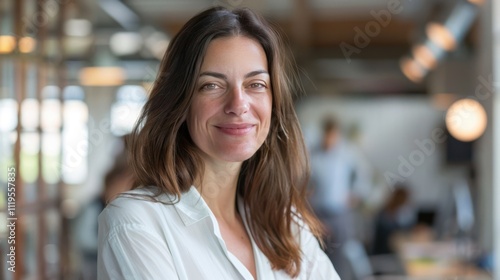 Smiling businesswoman in her 40s with long brown hair, wearing a white blouse, exuding confidence and professionalism in a modern, open office setting