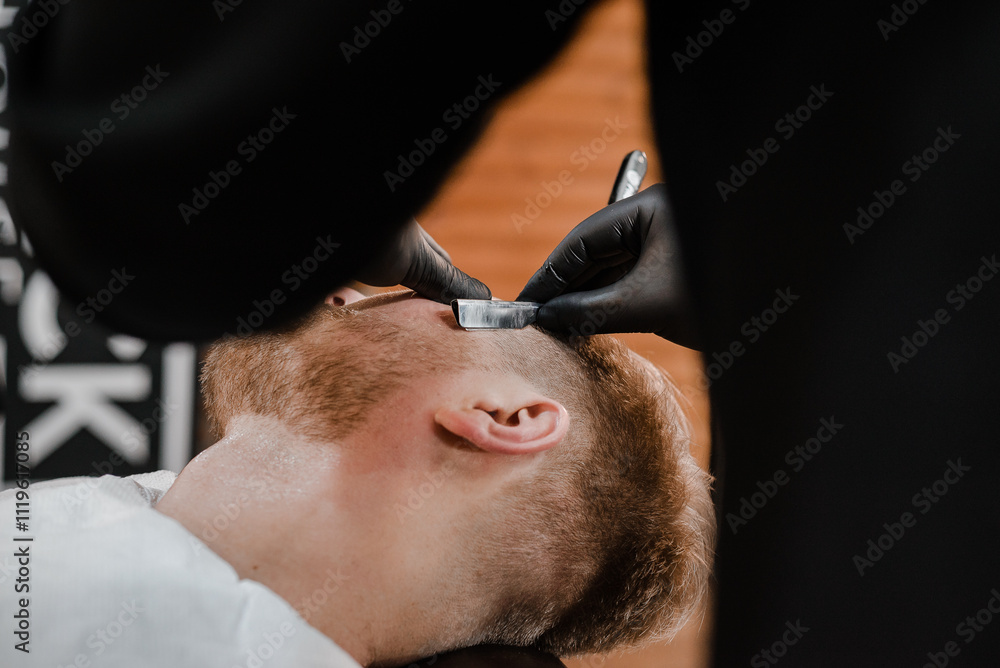 Fototapeta premium Barber shop. Man with a beard a client in a barbershop hairdresser Barber on a shaving with a dangerous razor.Shaving process,face closeup.