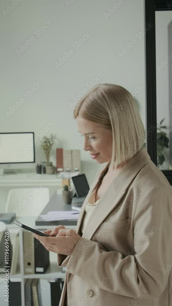 Vertical medium shot of smiling Caucasian businesswoman in stylish beige blazer standing in modern office setting with natural lighting while typing messages on her smartphone Vertical medium shot of smiling Caucasian businesswoman in stylish beige blazer standing in modern office setting with natural lighting while typing messages on her smartphone