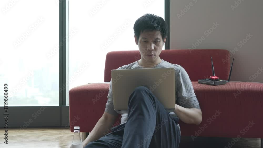 A man is sitting on a red couch with a laptop in front of him. He is focused on the screen, possibly working or browsing the internet. The couch and the laptop create a comfortable A man is sitting on a red couch with a laptop in front of him. He is focused on the screen, possibly working or browsing the internet. The couch and the laptop create a comfortable