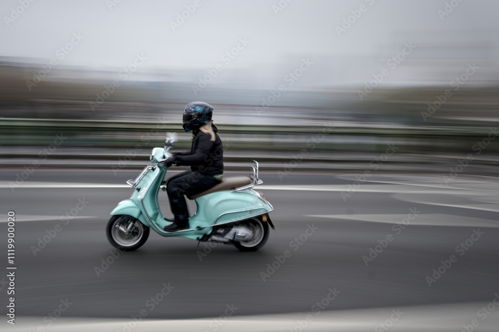 Blurred young woman on a green scooter, riding along a road to the left, with a panning effect. Blurred young woman on a green scooter, riding along a road to the left, with a panning effect.