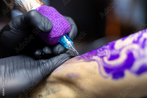 Close-up shot of a tattoo artist using a tattoo machine to create a detailed design on a client’s arm, with a focus on the purple stencil and black gloves. Perfect for tattoo art, creativity, and life