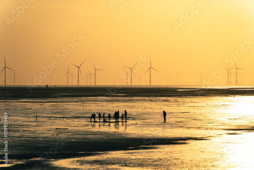 a group of people stand on a beach with a large wind farm in the background.