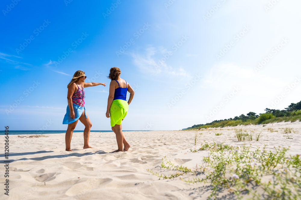Two mature women tourists standing and talking, one pointing something, on sandy beach in summer Two mature women tourists standing and talking, one pointing something, on sandy beach in summer
