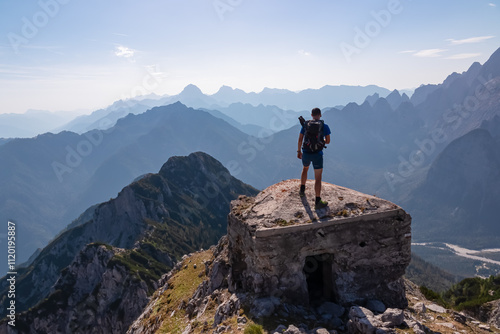 Hiker man on remains of first world war concrete bunker atop of mountain peak Jof di Miezegnot. Surrounded by majestic ridges of Julian Alps shrouded in soft, misty haze, Friuli Venezia Giulia, Italy photo