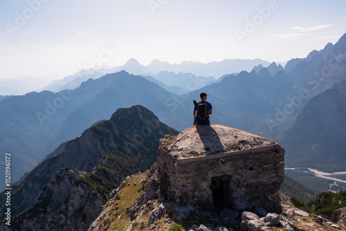 Hiker man on remains of first world war concrete bunker atop of mountain peak Jof di Miezegnot. Surrounded by majestic ridges of Julian Alps shrouded in soft, misty haze, Friuli Venezia Giulia, Italy photo