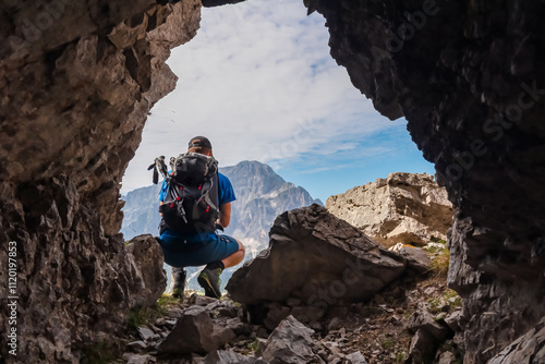 Hiker man in remnant of tunnel from first world war along defense line on mountain peak Jof di Miezegnot. Looking at majestic rugged massif Jof di Montasio, Julian Alps, Friuli Venezia Giulia, Italy photo