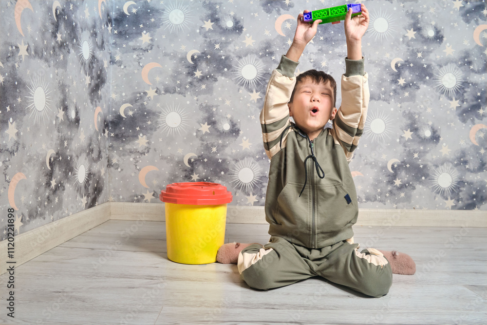 Naklejka premium A joyful boy in a tracksuit sits on the floor, holding a toy made of building blocks above his head
