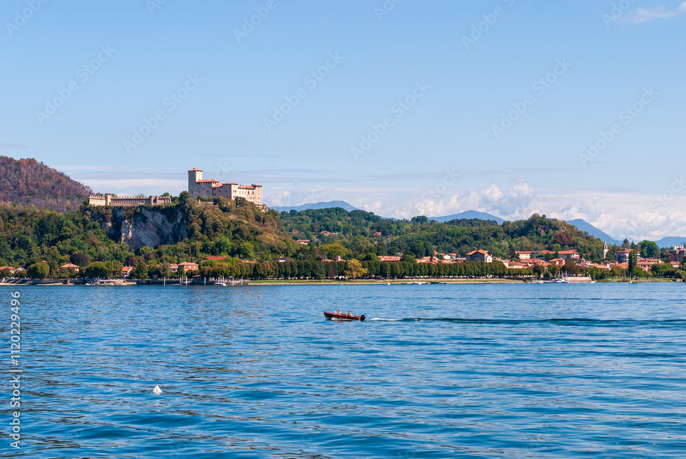 Fototapeta premium From Arona, the Rocca di Angera rises majestically above Lake Maggiore, its medieval fortress perched on a hill, offering a stunning contrast to the serene waters below.