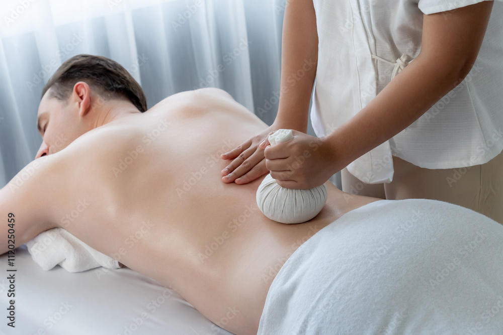 Fototapeta premium Hot herbal ball spa massage body treatment, masseur gently compresses herb bag on man body. Tranquil and serenity of aromatherapy recreation in day lighting ambient at spa salon. Quiescent