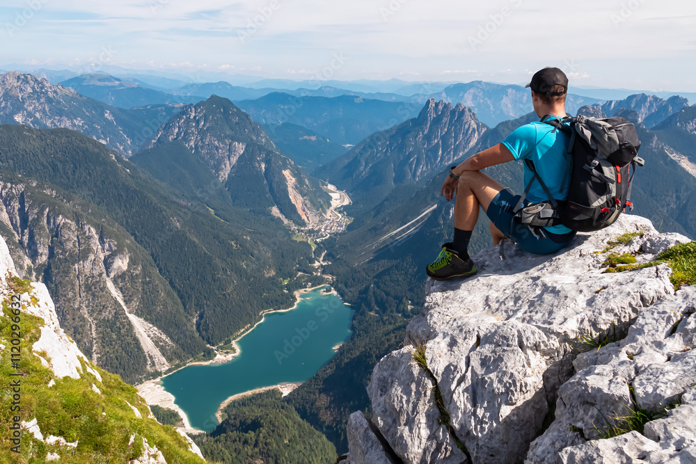 Naklejka premium Hiker man on top of Cima Del Lago with aerial view of alpine lake Lago Del Predil in Val Canale surrounded by majestic mountain peaks of Julian Alps, Friuli Venezia Giulia, Italy. Wanderlust in nature