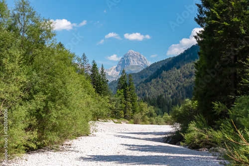 Scenic view of majestic mountain peak of Mangart in Julian Alps seen from Predil Pass, Tarvisio, Italy. Dried riverbed Rio Del Lago surrounded by pine tree forest. Hiking trail on alpine wilderness photo