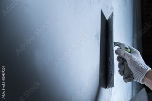 A hand with a glove is using a trowel to apply epoxy adhesive on a wall surface for repair. Microcement or decorative plaster.