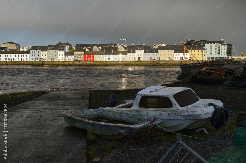 Naklejka premium Beautiful cityscape sunrise scenery with old wooden boat in Corrib river at Claddagh in Galway city, Ireland, architecture background