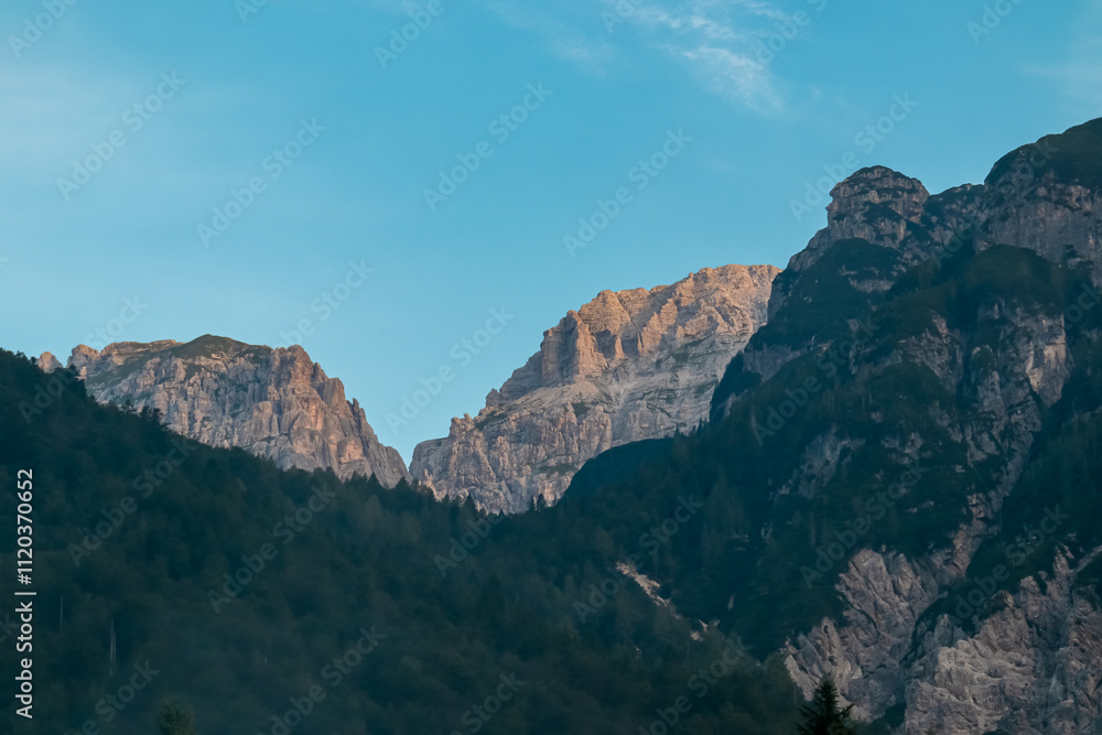 Scenic view of majestic mountain peak of Mangart in Julian Alps seen from Predil Pass, Tarvisio, Italy. Ridges covered in morning mist. Hiking trail on alpine terrain covered with conifer forest Scenic view of majestic mountain peak of Mangart in Julian Alps seen from Predil Pass, Tarvisio, Italy. Ridges covered in morning mist. Hiking trail on alpine terrain covered with conifer forest