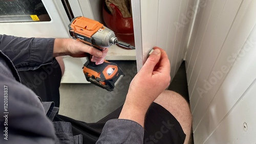 A handyman is using a power drill to replace the knob on a wooden kitchen cupboard. The setting is a clean, white kitchen with the cupboard door slightly open, revealing a glimpse of storage inside. 