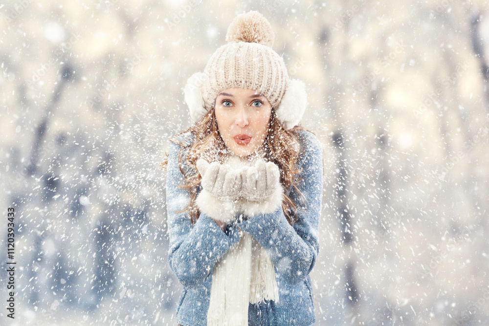 Happy young woman blowing snow from her hands looking into the camera, outdoors during falling snow. Christmas holidays or winter season vacation and cooling and heating temperatures concept Happy young woman blowing snow from her hands looking into the camera, outdoors during falling snow. Christmas holidays or winter season vacation and cooling and heating temperatures concept